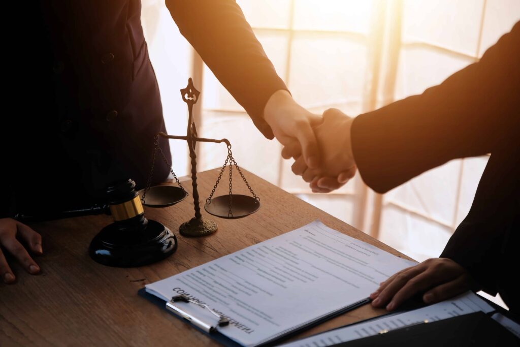 Lawyers shaking hands over a signed legal agreement with a judge&rsquo;s gavel and justice scales on the desk, representing legal consultation and contract settlement.