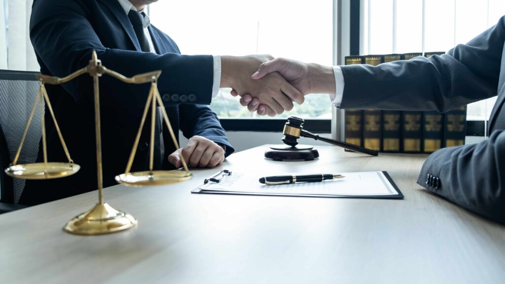 Two lawyers shaking hands across a desk with justice scales, a judge&rsquo;s gavel, and legal documents, symbolizing a law firm agreement and case settlement