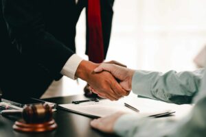 Lawyer and client shaking hands over a signed contract with a judge&rsquo;s gavel on the desk, symbolizing legal agreement and case settlement.
